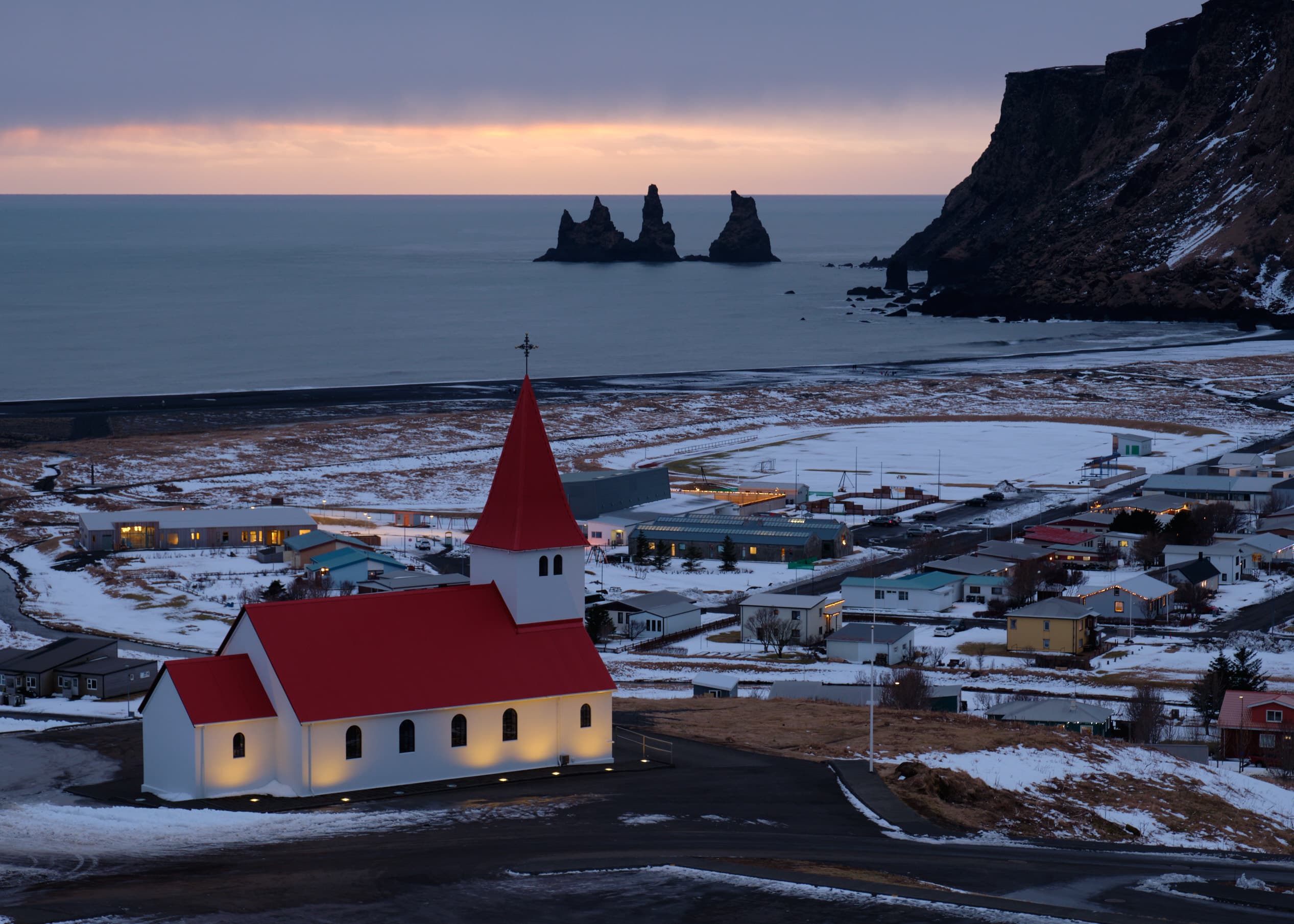 Vík church lit at dusk, sea stacks and village below